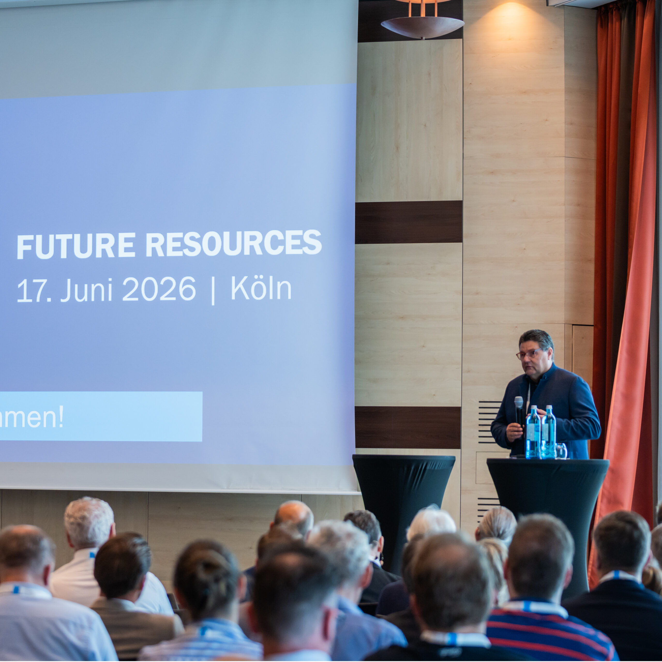Audience seated in a conference hall facing a speaker at a lectern with water bottles; a blue banner reads 'FUTURE RESOURCES'.