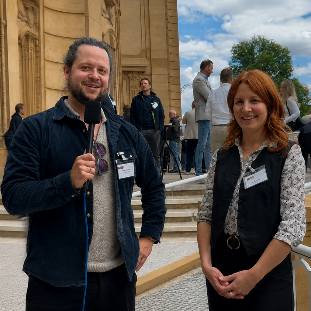Man with a microphone and name badge speaks at an outdoor event beside a smiling woman with red hair and a vest, architectural backdrop behind them.